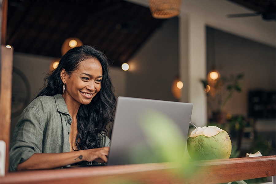 A smiling woman works on her laptop at a tropical location, with a coconut drink beside her.